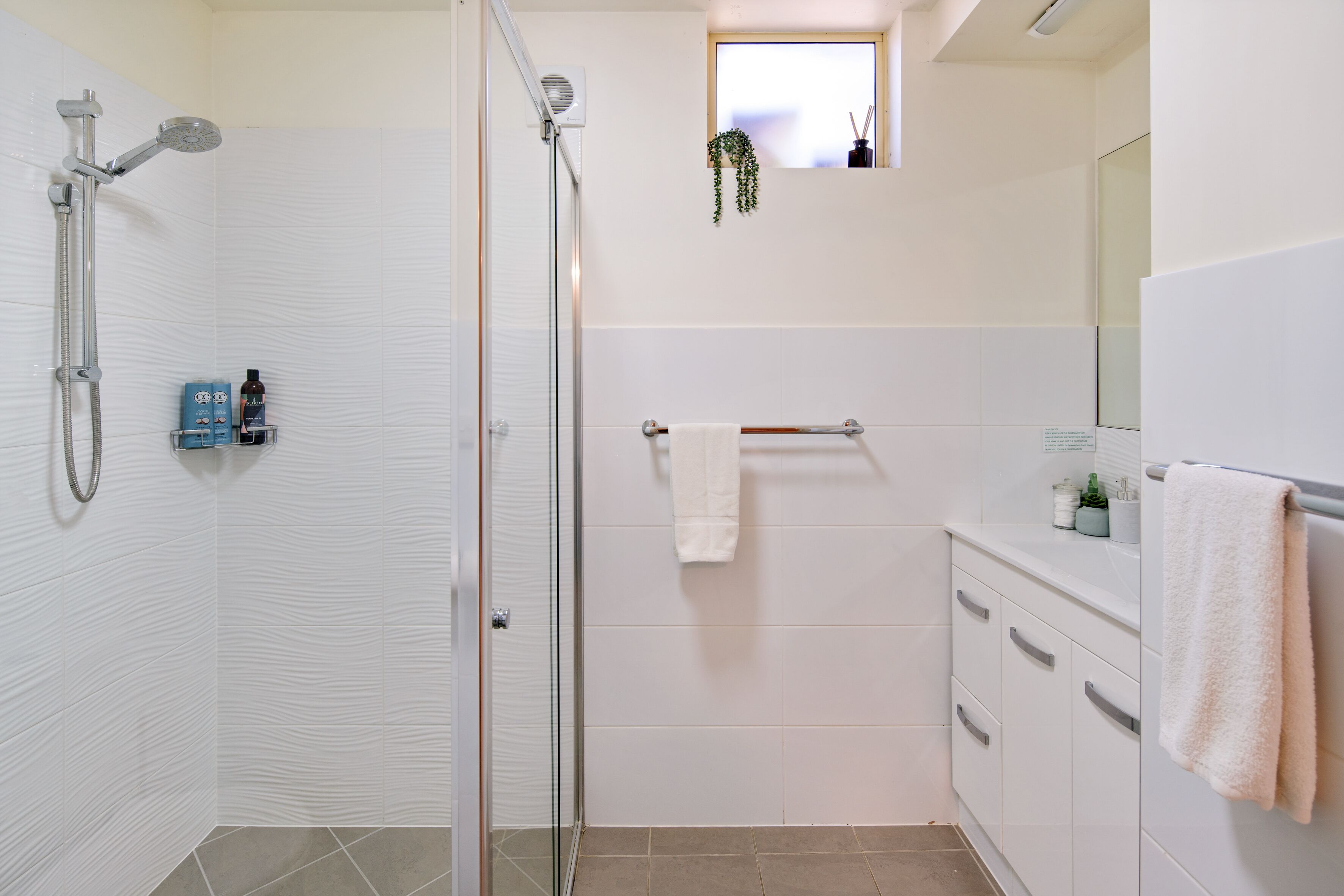 Un baño moderno con cabina de ducha de vidrio, azulejos blancos, un ventanal con una planta y un mueble de tocador con toallero.