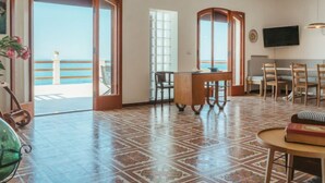 A spacious room with a large open door leading to a balcony, a dining area with wooden furniture, and a television mounted on the wall. - Santa Maria by Interhome (Marina di Caronia)