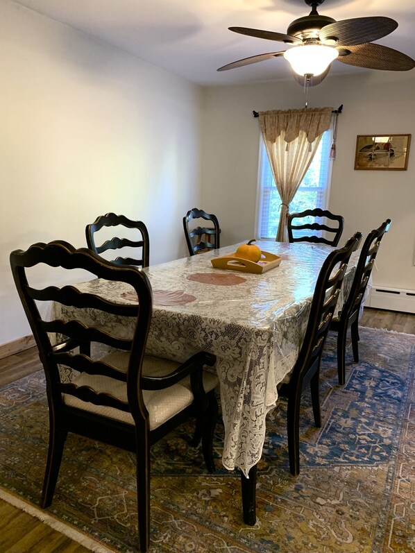 Un comedor con una mesa cubierta por un mantel de encaje, rodeada de sillas de madera con asientos acolchados. Se observa un ventilador de techo sobre la mesa, y una ventana con cortinas en la pared.