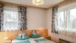 Une chambre à coucher comprenant un lit, deux tables de chevet, une commode, une fenêtre avec des rideaux à motifs floraux et un luminaire au plafond.