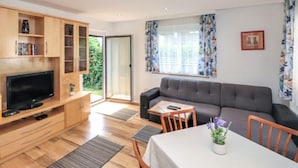 Un salon moderne doté d’un téléviseur à écran plat, de meubles en bois, d’un canapé gris, d’une table à manger avec des chaises et d’une grande fenêtre agrémentée de rideaux à motifs floraux.