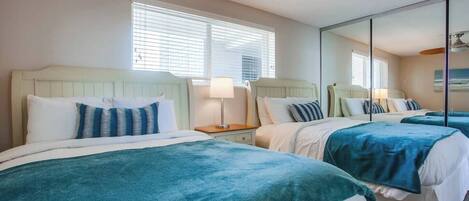 A hotel room with two beds, a ceiling fan, a bedside table with a lamp, and a large mirror.