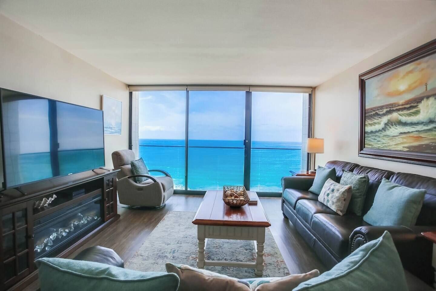 A living room with a large TV, a sofa, a coffee table, and a view of the ocean through large windows.