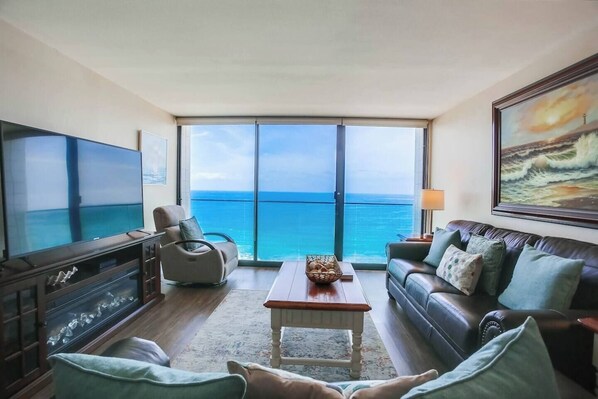 A living room with a large TV, a sofa, a coffee table, and a view of the ocean through large windows.