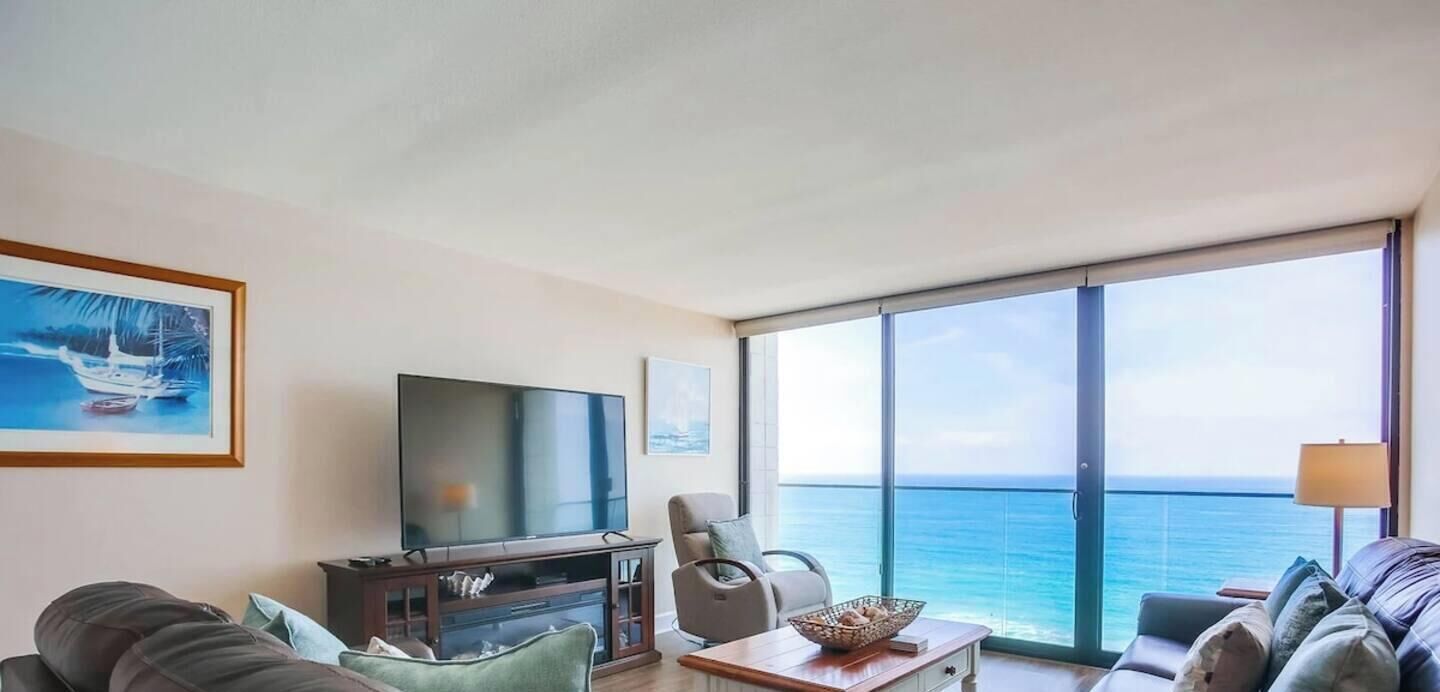 A living room with a large flat-screen TV, a sofa, a chair, and a table with a decorative bowl. There is a large window offering a view of the ocean.