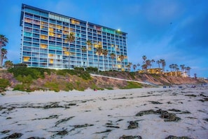 Un hôtel moderne à l’architecture imposante, avec plusieurs étages, situé à proximité d’une plage bordée de palmiers et d’une côte rocheuse.