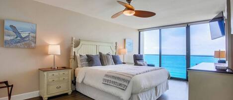 Un dormitorio con cama, mesitas de noche, ventilador de techo, un amplio ventanal con vista al mar y obras de arte en la pared.