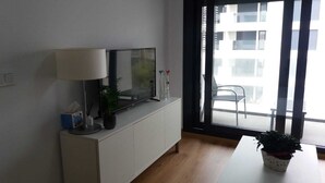 A modern living room with a TV on a white cabinet, a lamp, a potted plant, and a view of a balcony with a chair.
