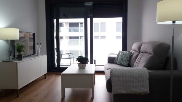 A modern living room with a sofa, a coffee table, and a TV on a stand. There is a window with a view of buildings outside.