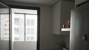 A compact kitchen with a built-in oven, a refrigerator, and a window with blinds.