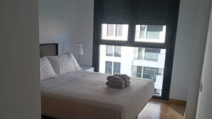 A bedroom with a bed, a nightstand, and a window with a view of buildings.