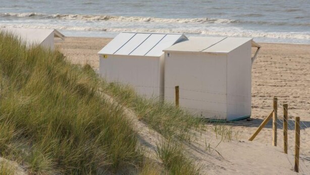 Une plage avec deux cabanes de plage blanches et un voilier à l’horizon.