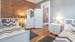 Une chambre avec un mur en briques, deux lits, des meubles blancs et une porte donnant sur une autre pièce.