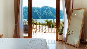 Un dormitorio con una cama grande, un espejo con marco de madera y un balcón con vistas a una colina boscosa y un lago.