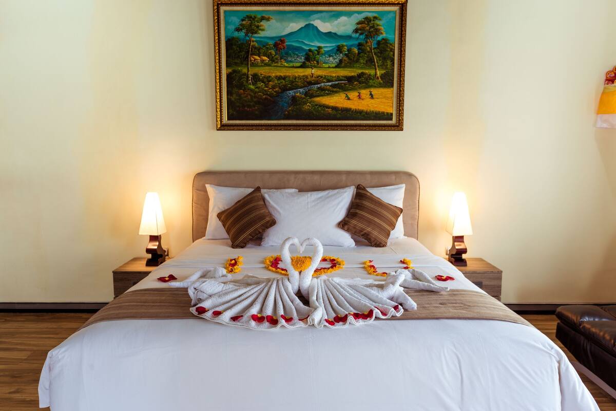 A neatly made bed with a towel arrangement in the shape of swans, a painting on the wall, and two bedside lamps.