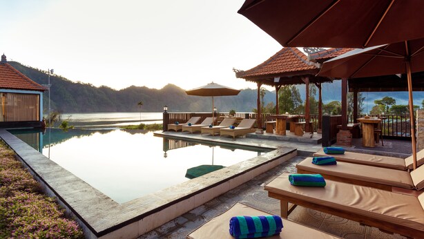 Zona de piscina con sillas de descanso y sombrillas, rodeada por una terraza de madera y con vista a una masa de agua.
