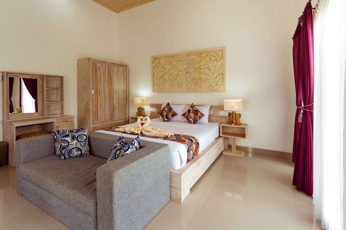 A bedroom with a bed, two bedside tables, a wooden wardrobe, a grey sofa, and a large wall art piece.