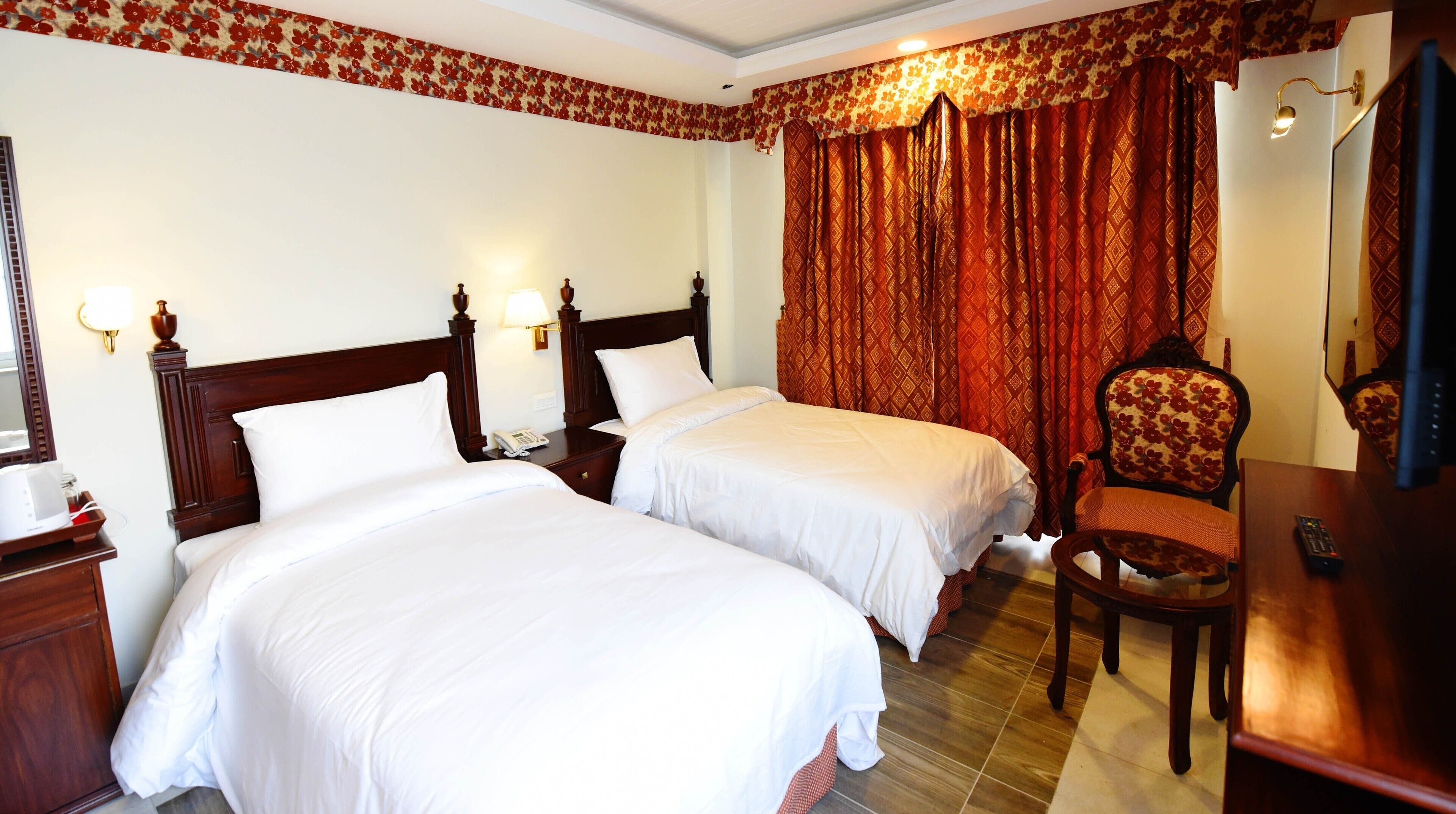A hotel room with two single beds, a wooden nightstand with a lamp, a chair, and a desk with a television and remote control.
