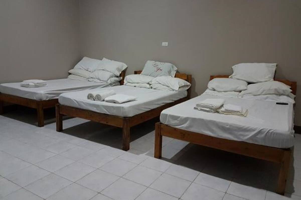 Three single beds with wooden frames and white bedding, arranged in a row in a room with plain walls and tiled flooring.