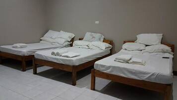 Three single beds with wooden frames and white bedding, arranged in a row in a room with plain walls and tiled flooring.