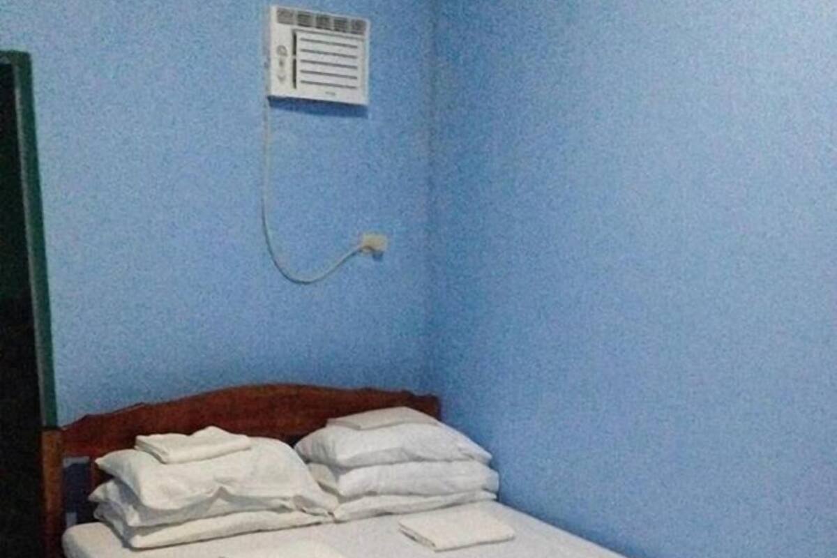A single bed with white linens and pillows, a power outlet on the wall, and a tiled floor.