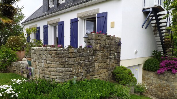 A house with a stone wall, blue shutters, and a garden with various plants and flowers. - Independent dwelling in 1 detached house near sea. (GUIDEL)