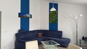A modern living room with a blue sectional sofa, a glass coffee table, and a white pendant light. - Renovated vacation apartment Mare - Holiday apartment Mare in Zinnowitz (Zinnowitz)