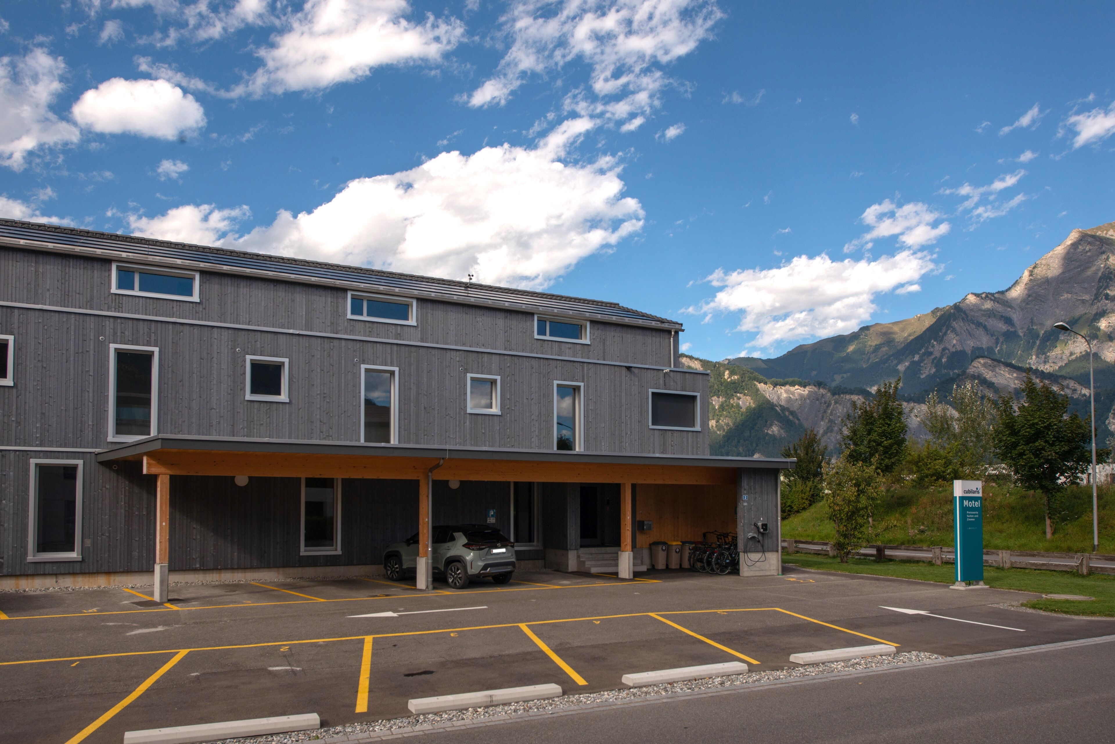 Un bâtiment moderne doté d’un parking couvert, d’une voiture et ayant une chaîne de montagnes en arrière-plan.