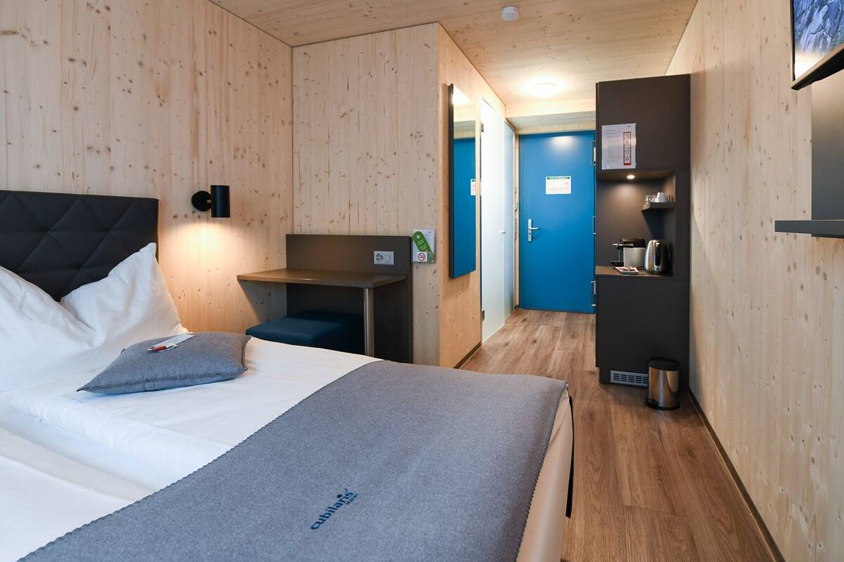 A hotel room with a bed, a desk, a blue door, and a kitchenette.