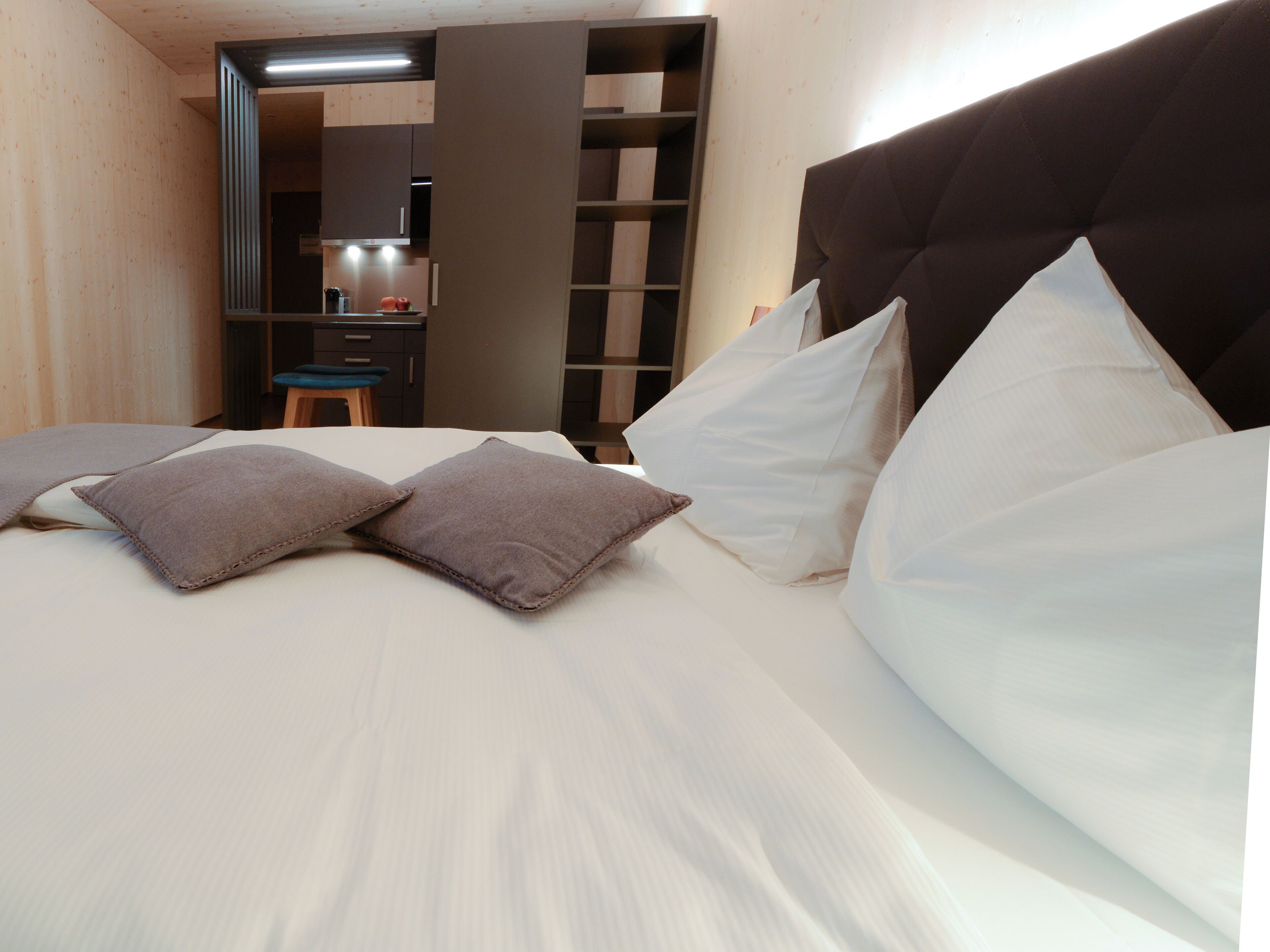 A bed with white and gray pillows, a wooden headboard, and a view of a kitchen area with a stool and cabinets.