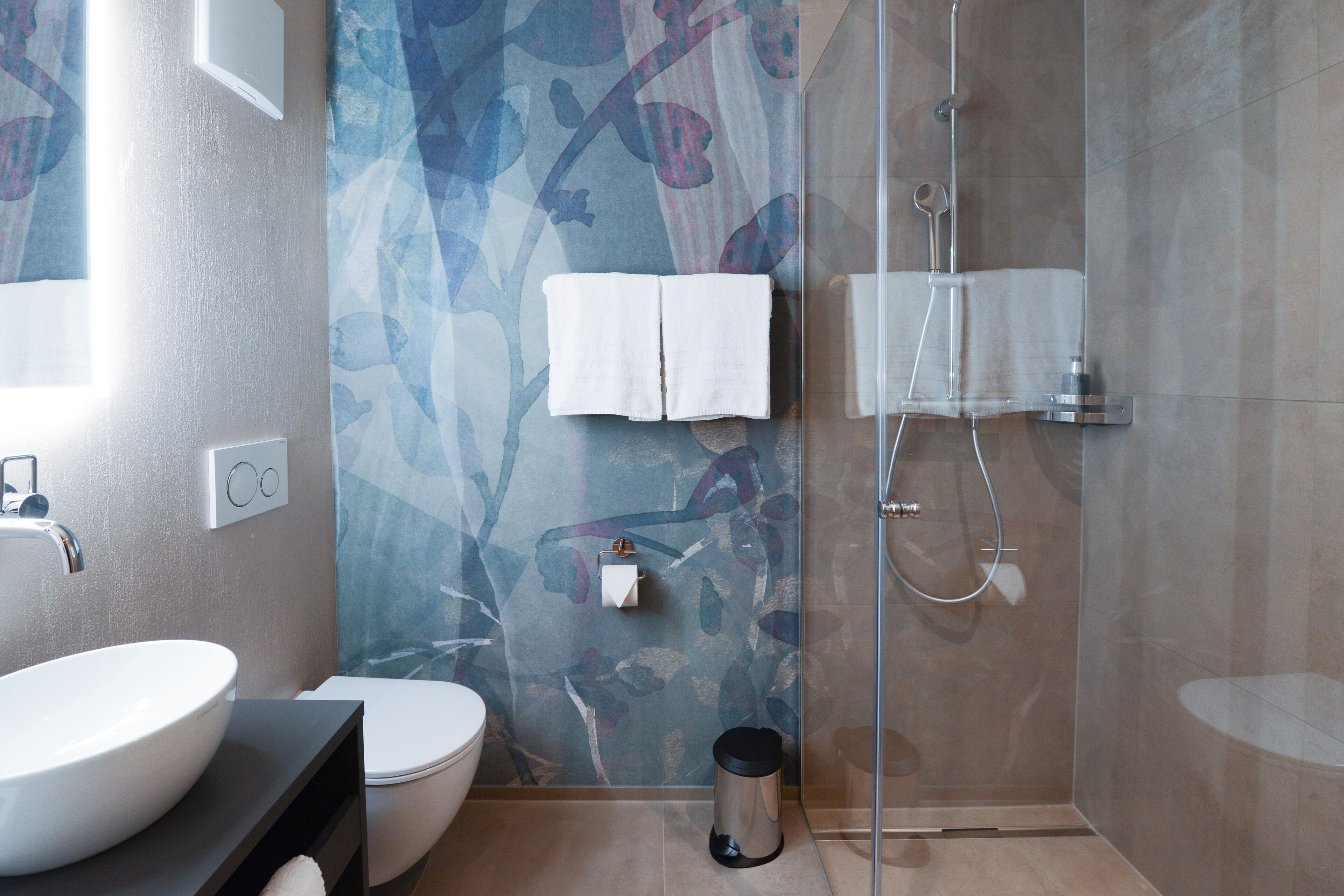 Un baño moderno con un lavabo blanco grande, un inodoro, una cabina de ducha de vidrio y un revestimiento de pared estampado.