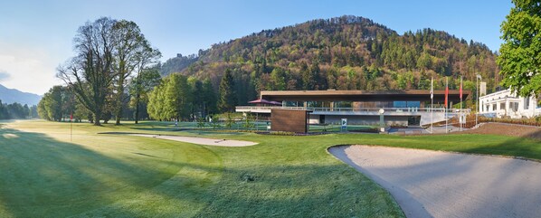 A golf course with a modern clubhouse, a mountain backdrop, and a clear sky.