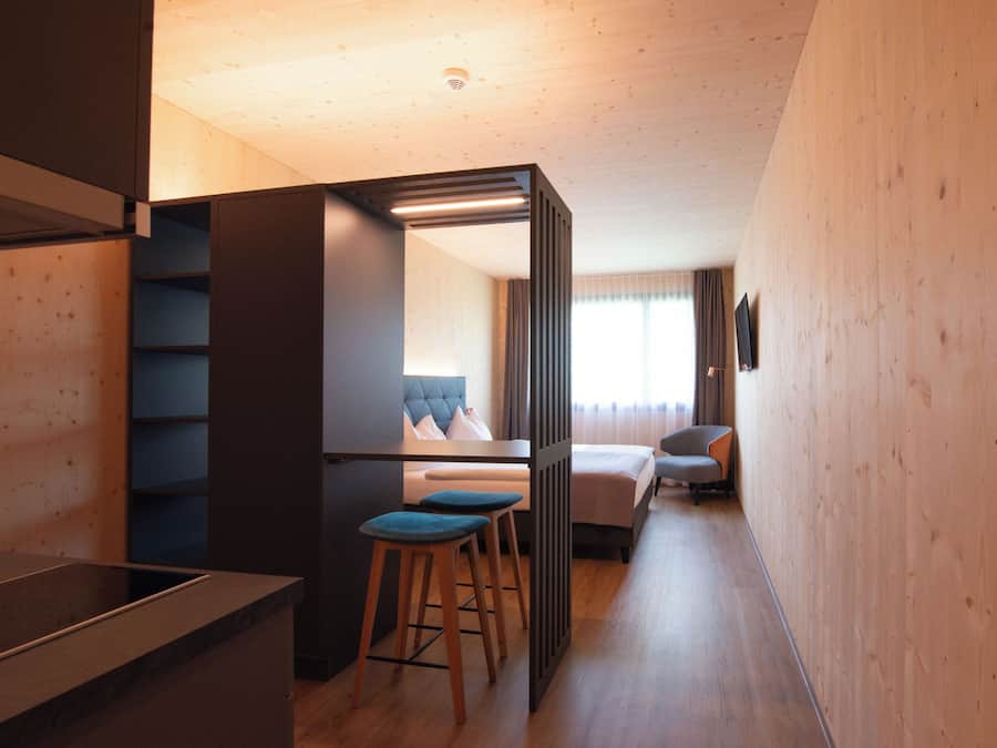 A modern hotel room with a wooden interior, a bed, a desk, and two bar stools.
