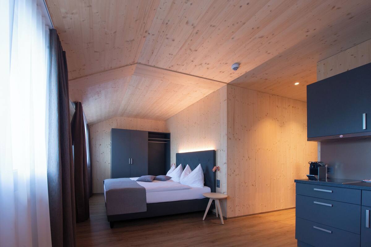 A modern hotel room with a wooden ceiling, a bed with white bedding, a small bedside table, and a built-in wardrobe.