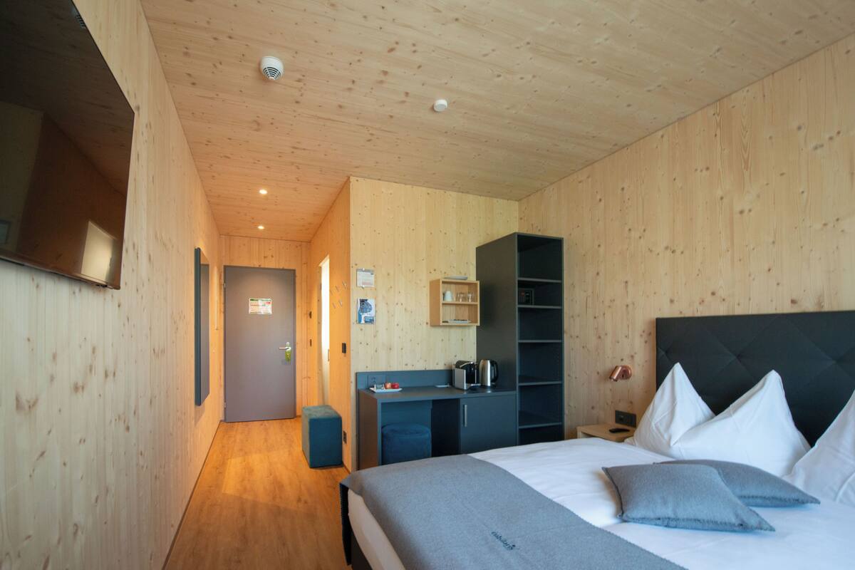 A modern hotel room with a wooden interior, a bed with pillows, a desk with a kettle, and a wardrobe.