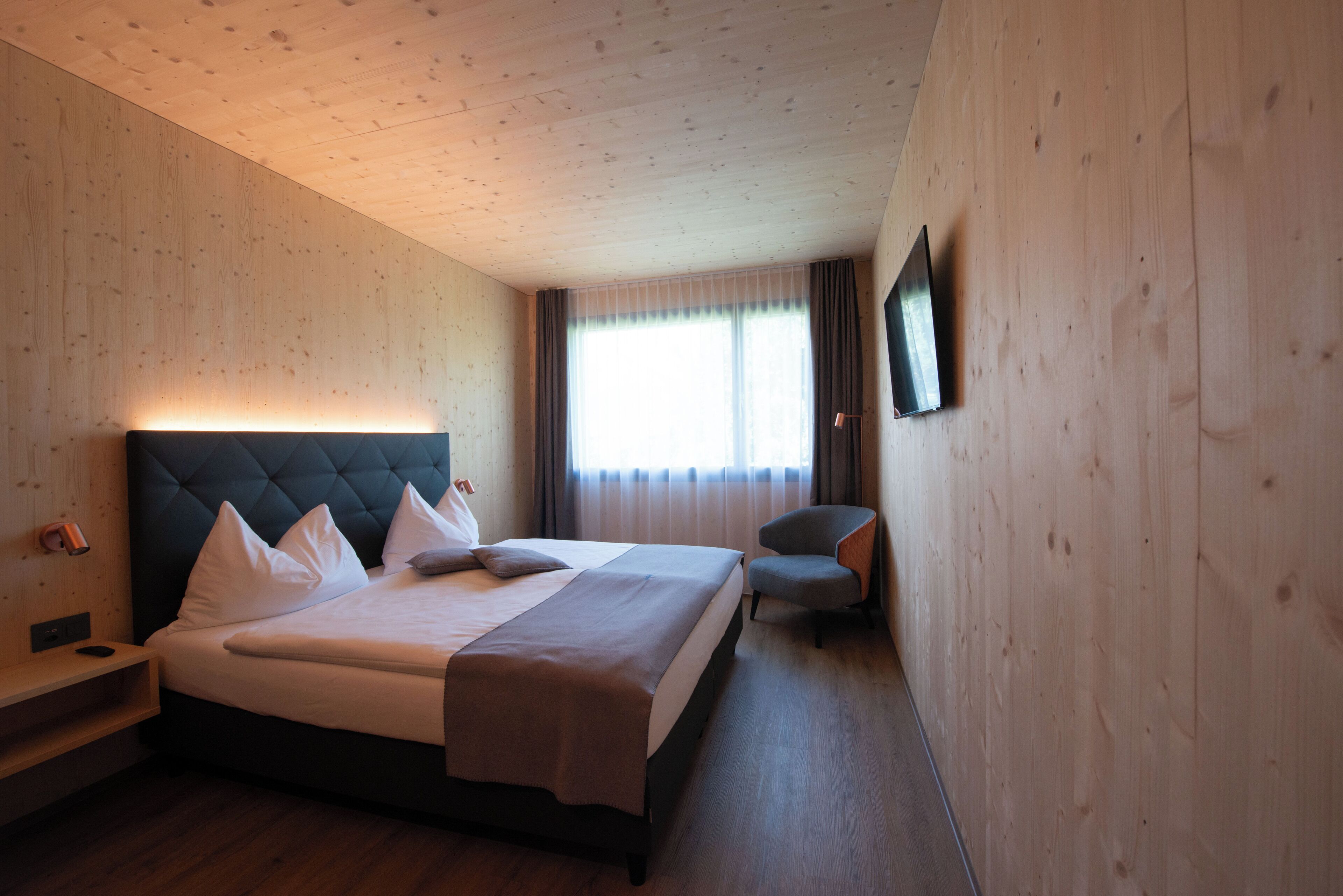 Ein Hotelzimmer mit einem großen Bett, einem Nachttisch, einem Sessel, einem an der Wand montierten Fernseher und einem fenster mit Vorhängen.