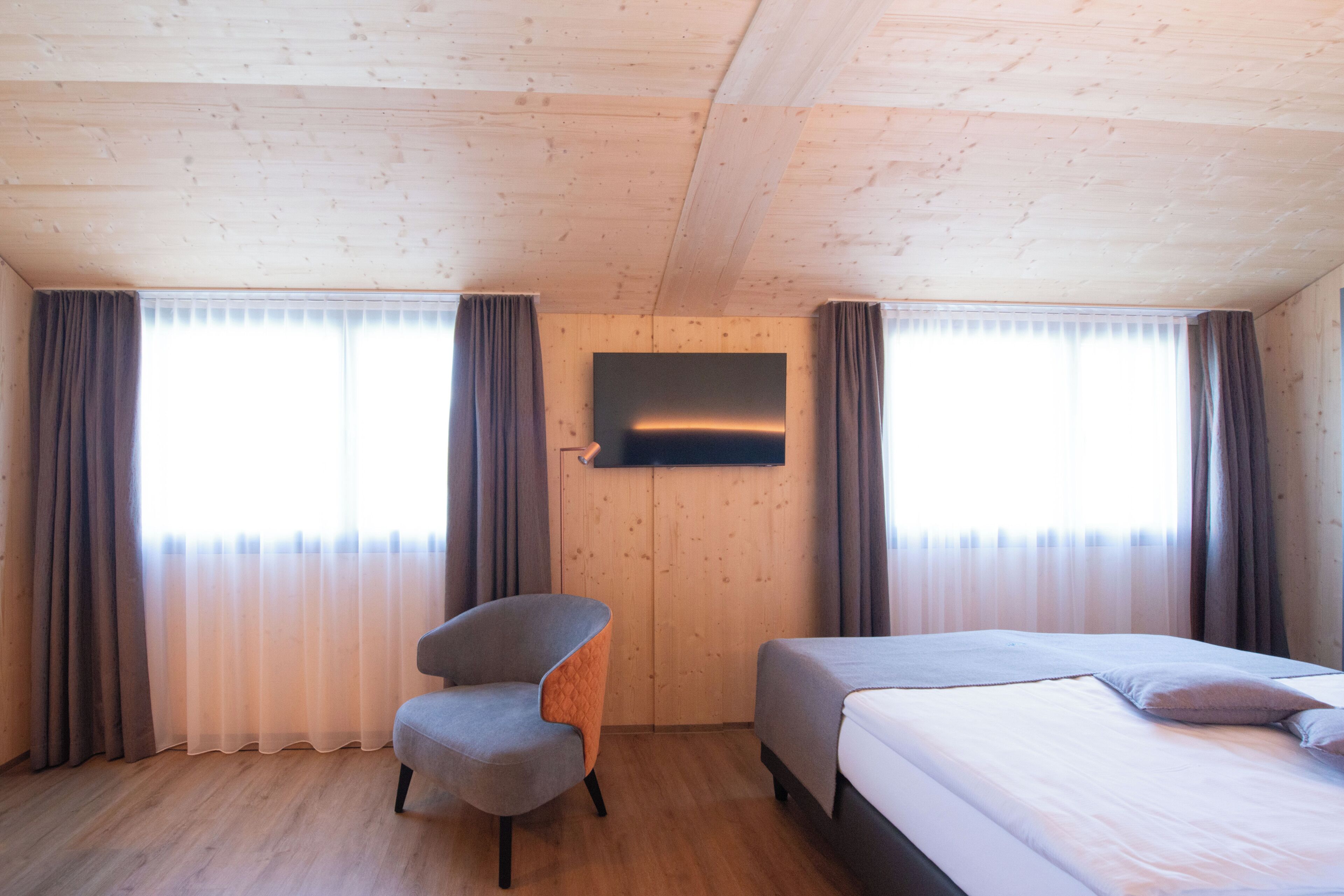 A room with a wooden ceiling, a flat-screen TV mounted on the wall, a grey armchair, and a bed with a white mattress and grey bedding.