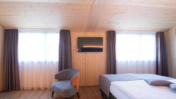 A room with a wooden ceiling, a flat-screen TV mounted on the wall, a grey armchair, and a bed with a white mattress and grey bedding.