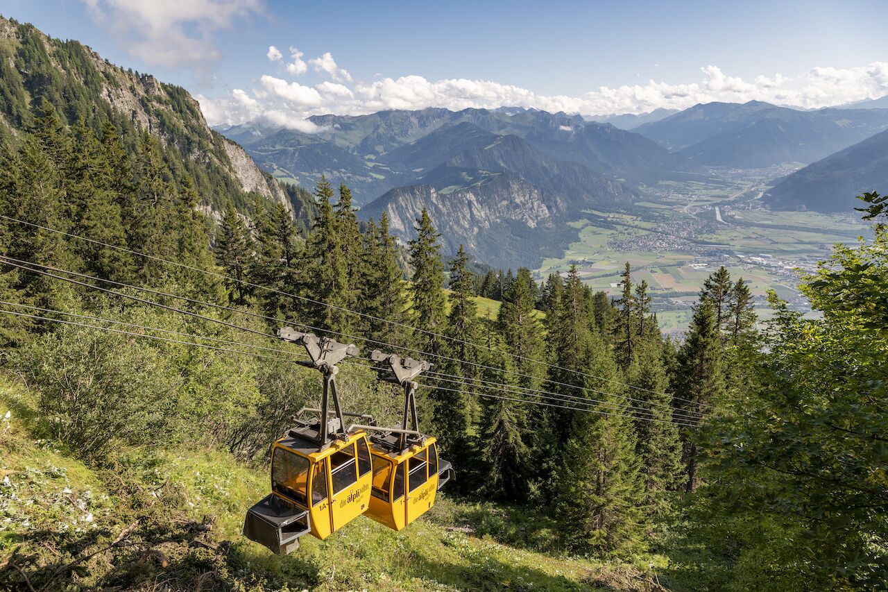 Sistema de teleférico en zona montañosa con abundante vegetación y vistas a una población distante.