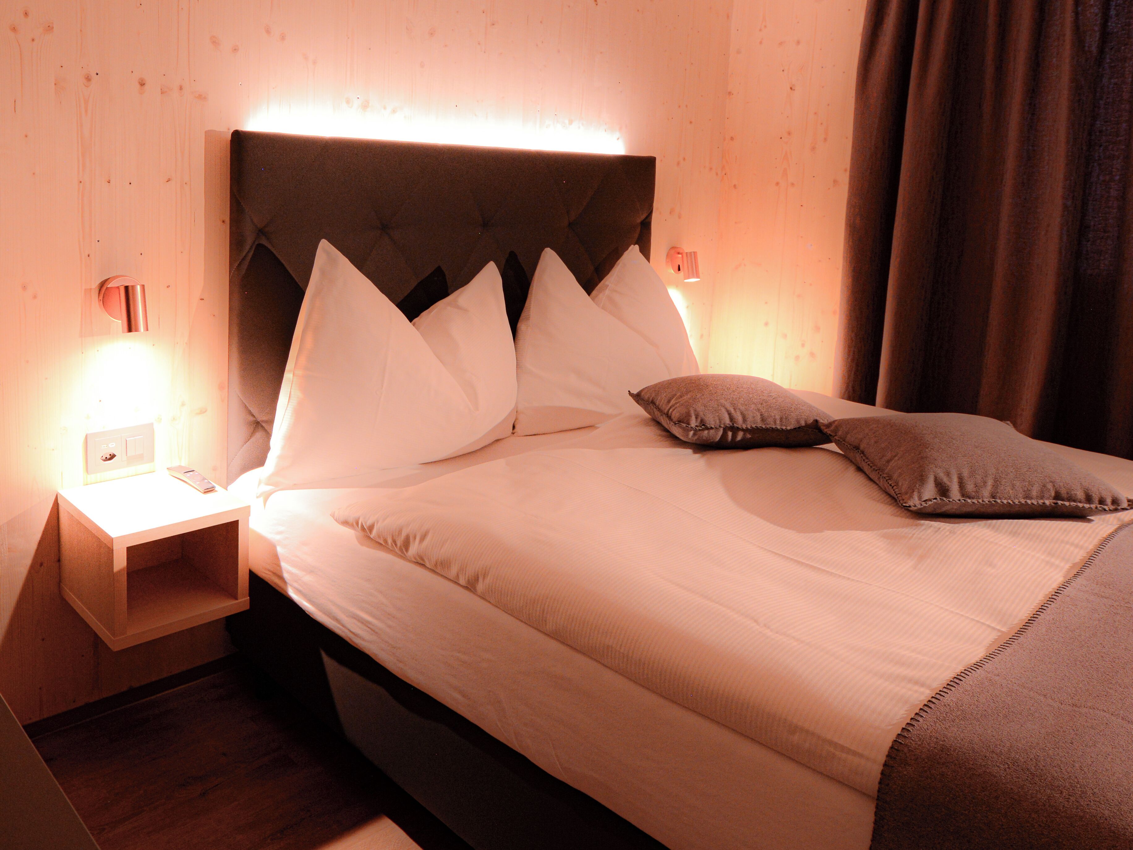 Una cama bien hecha con sábanas blancas y un cabecero marrón, junto a dos lámparas de noche y una mesita de noche de madera.