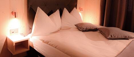 Una cama bien hecha con sábanas blancas y un cabecero marrón, junto a dos lámparas de noche y una mesita de noche de madera.