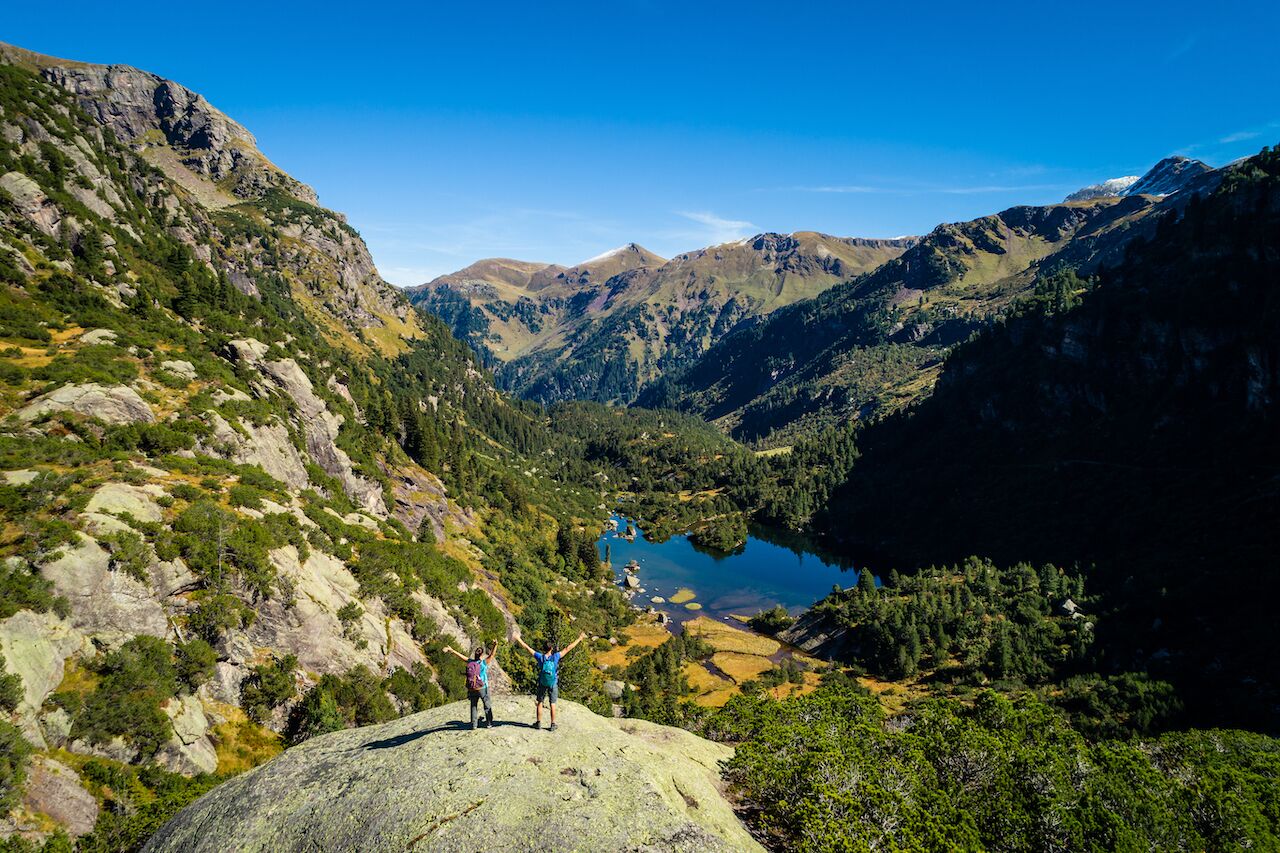 A mountainous landscape with a lake and two people standing on a rock.