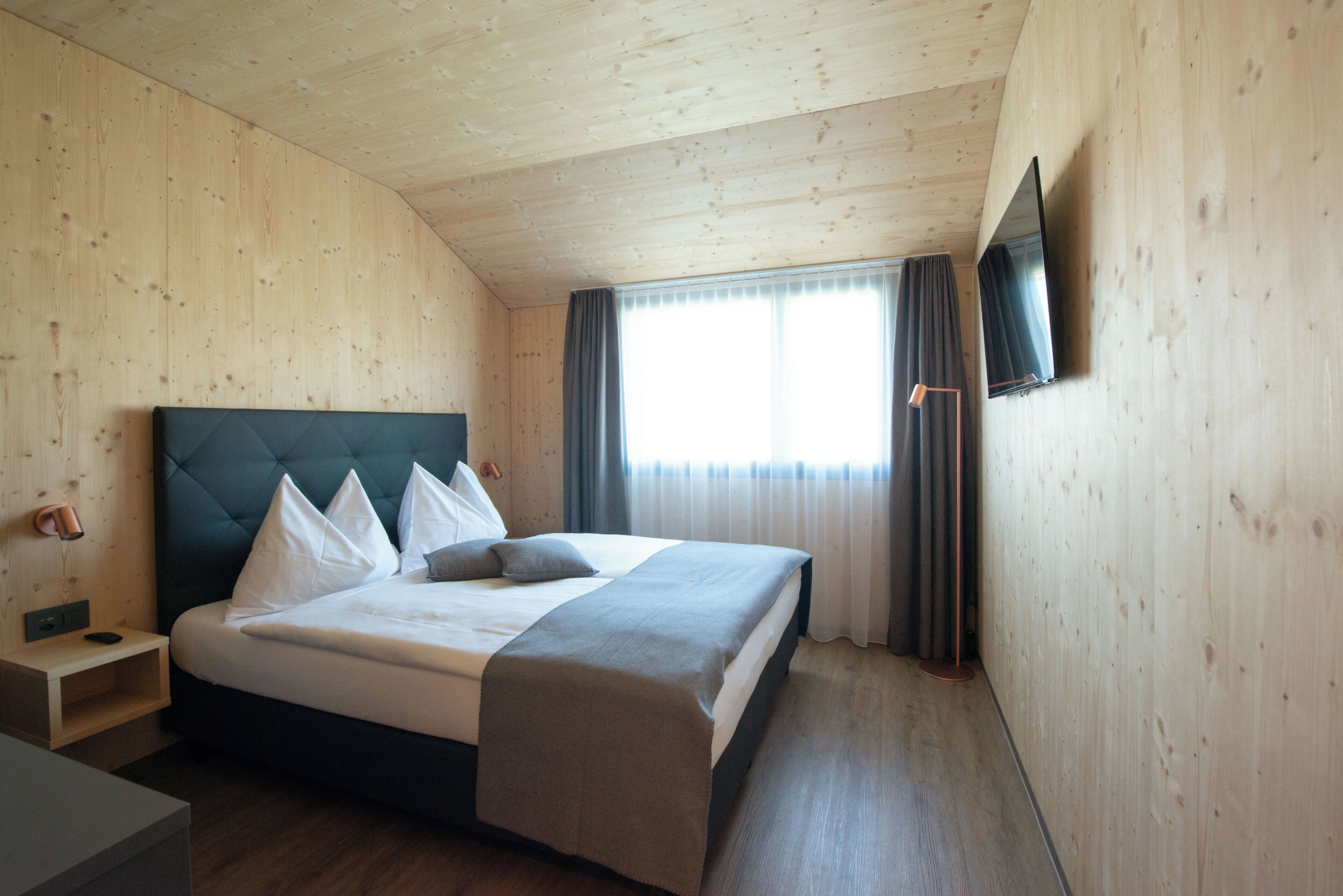 Ein Schlafzimmer mit Holzboden, einem Bett mit weißer Bettwäsche, einem Nachttisch, einem an der Wand montierten Fernseher und einem Fenster mit Vorhängen.