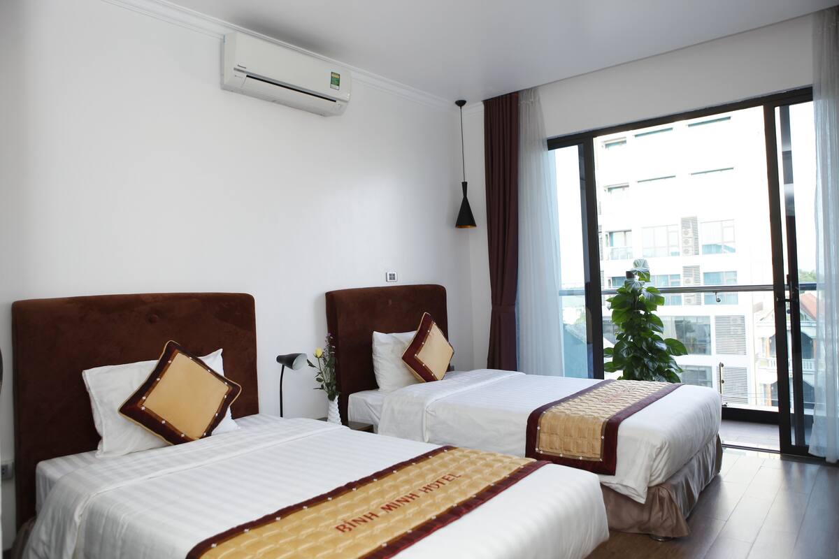 Ett hotellrum med två sängar, ett stort fönster med utsikt över byggnader och en luftkonditioneringsanläggning på väggen.