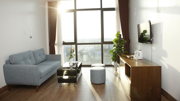 A modern living room with a grey sofa, a wooden coffee table, a white ottoman, and a wooden side table with a plant and a kettle. - Binh Minh Hotel (Dong Van)