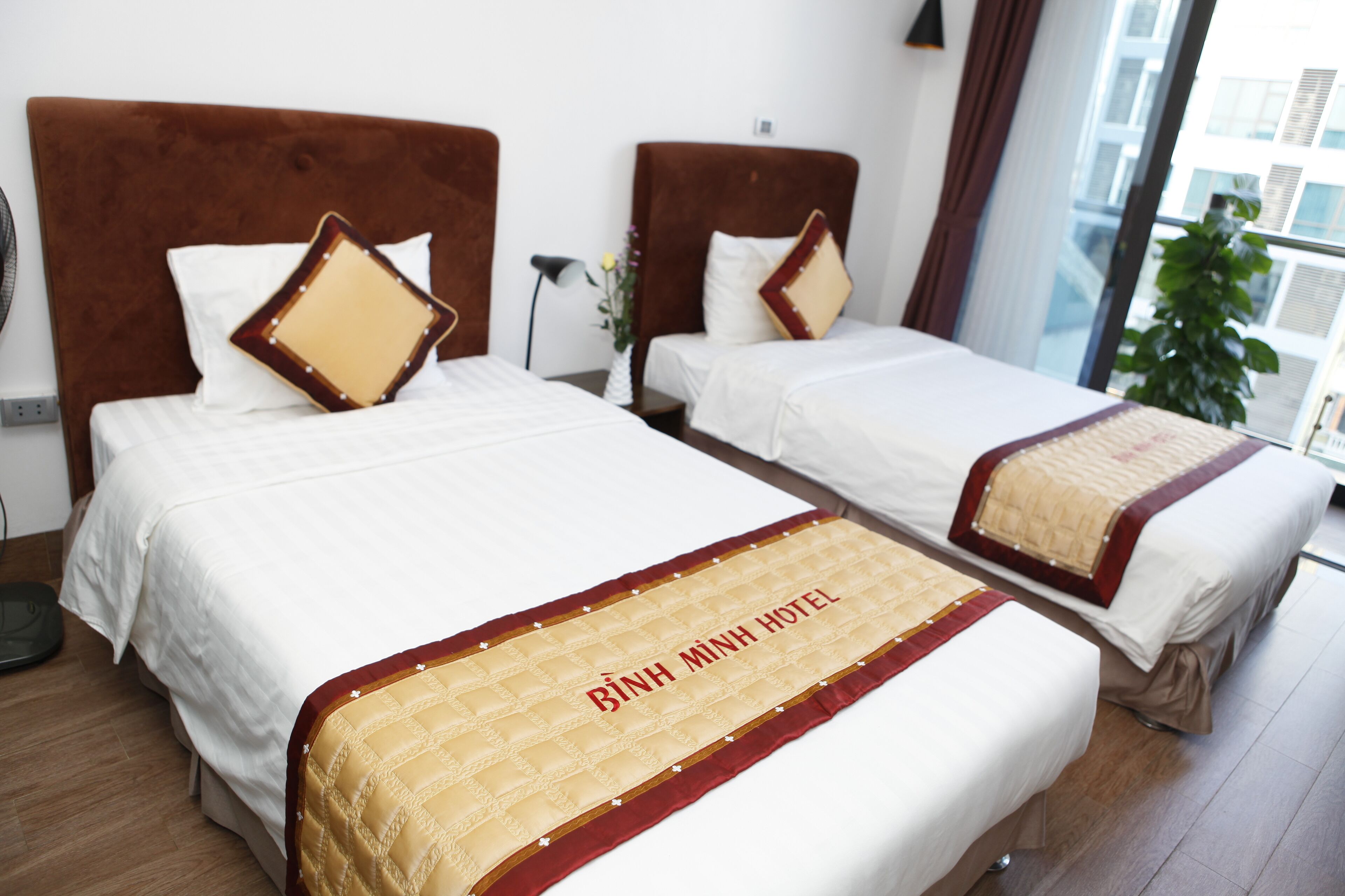 Two twin beds with headboards, a bedside table, a lamp, and a window with a view of buildings.