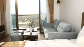 A hotel room with a bed, a grey sofa, a small table with a vase, and a view of the city through large windows. - Binh Minh Hotel (Dong Van)