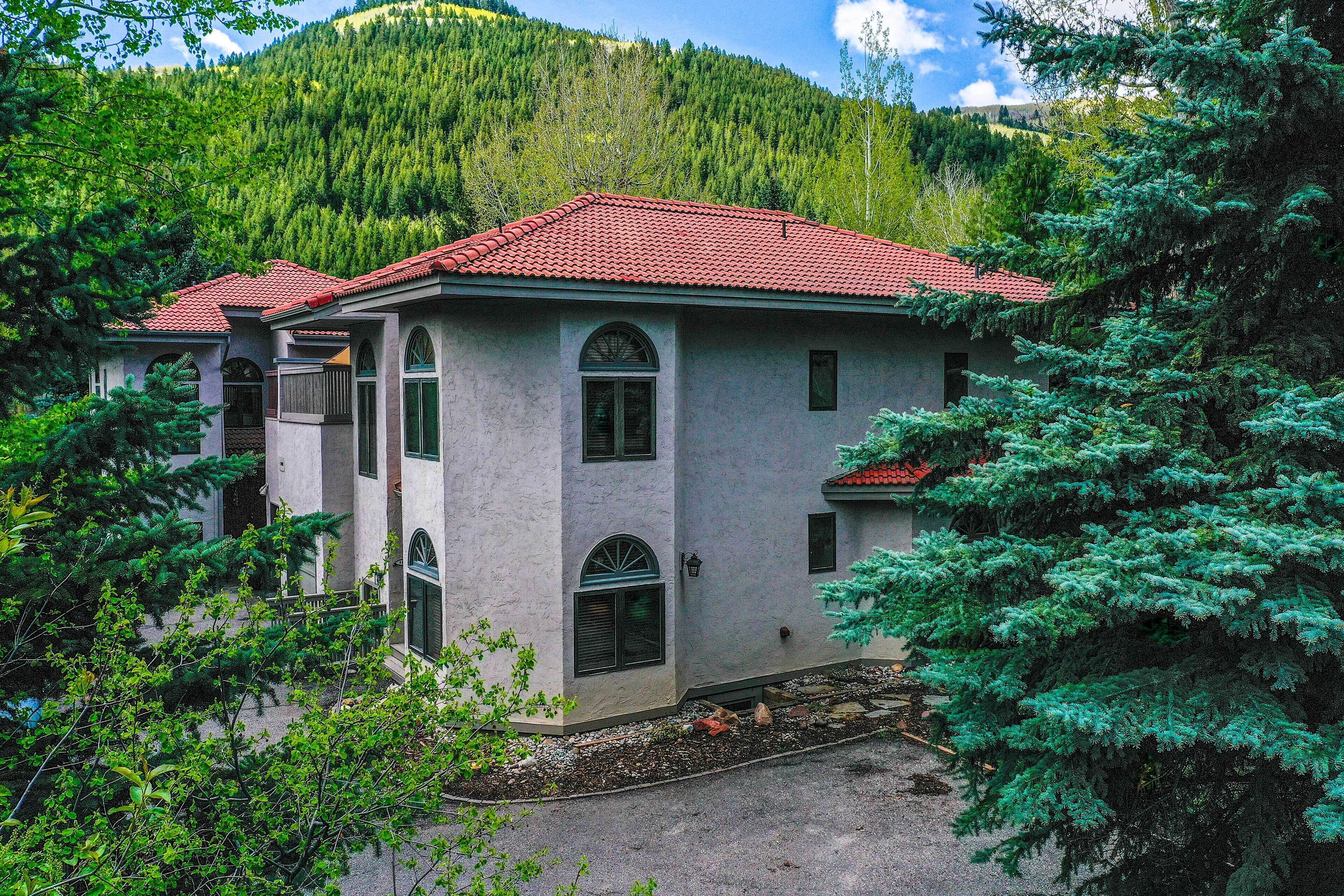 A two-story house with a red-tiled roof, surrounded by lush greenery and a mountainous backdrop.