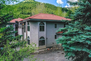 Una casa de dos pisos con tejado de tejas rojas, rodeada de abundante vegetación y montañas al fondo.