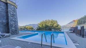 A rectangular outdoor swimming pool with metal ladders, surrounded by lounge chairs, a stone wall, and a view of mountains. - Palma Romantic Dream (Perledo)